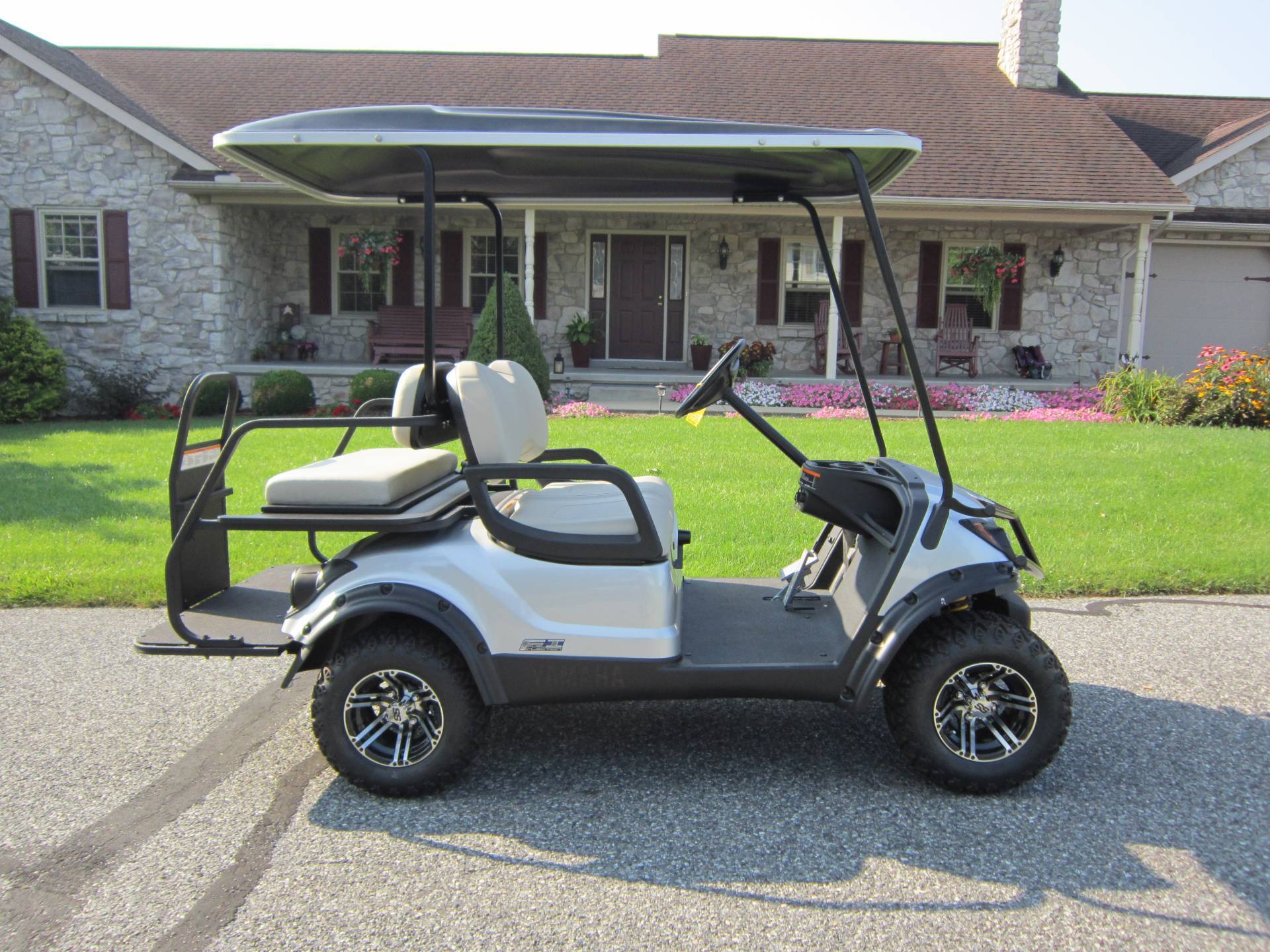 A sleek Burkholder Golf Cart with black accents is parked on a paved surface in front of a stone house. It features two seats, a roof, and large wheels. A well-manicured lawn and vibrant flower beds enhance the charming scene, complemented by a porch with red shutters.