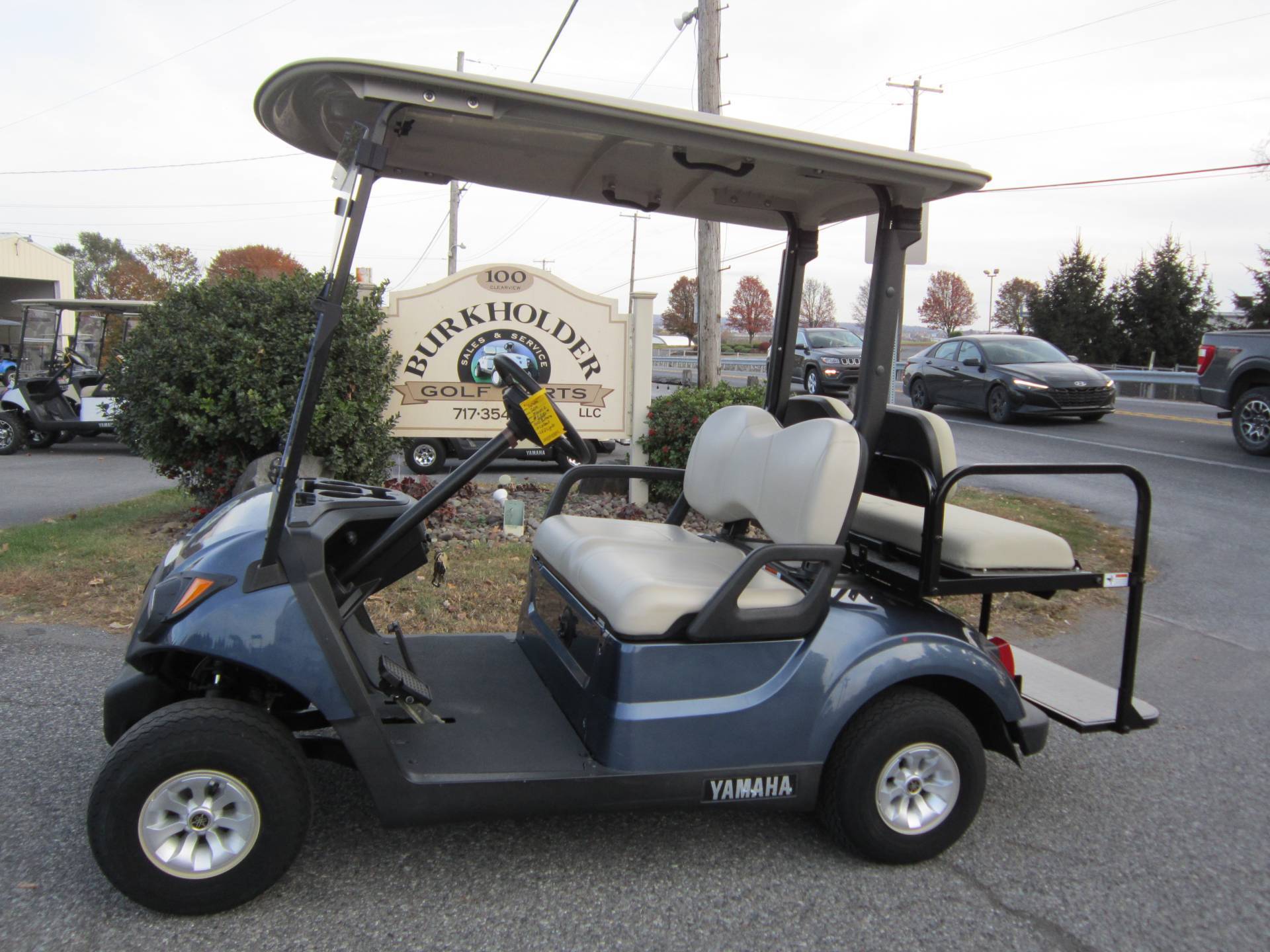 A blue Yamaha golf cart with beige seats parked outside Burkholder Golf Carts sign, surrounded by autumn foliage.